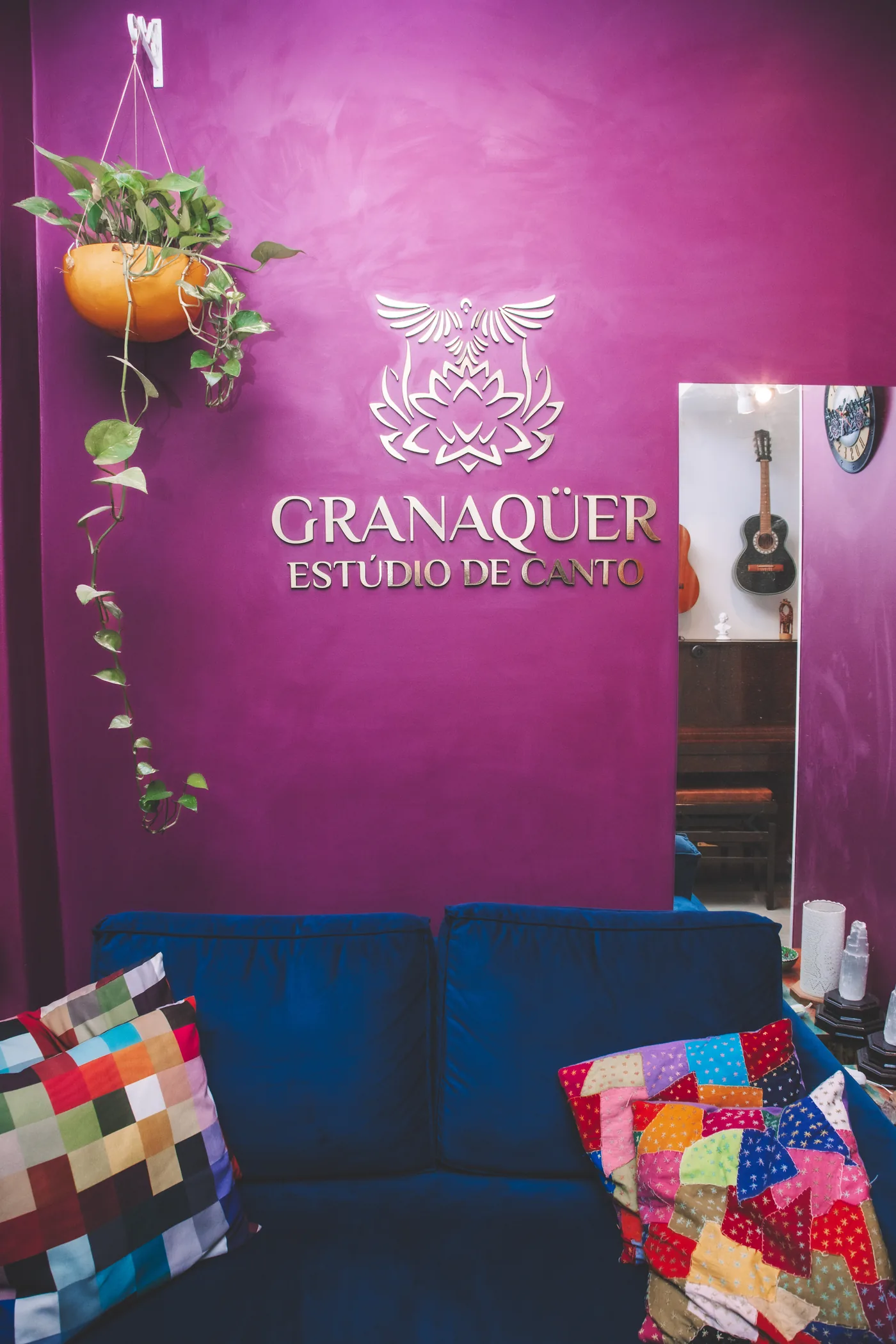 Parede violeta com o logotipo dourado Granaqüer Estúdio de Canto sobre o sofá da recepção
