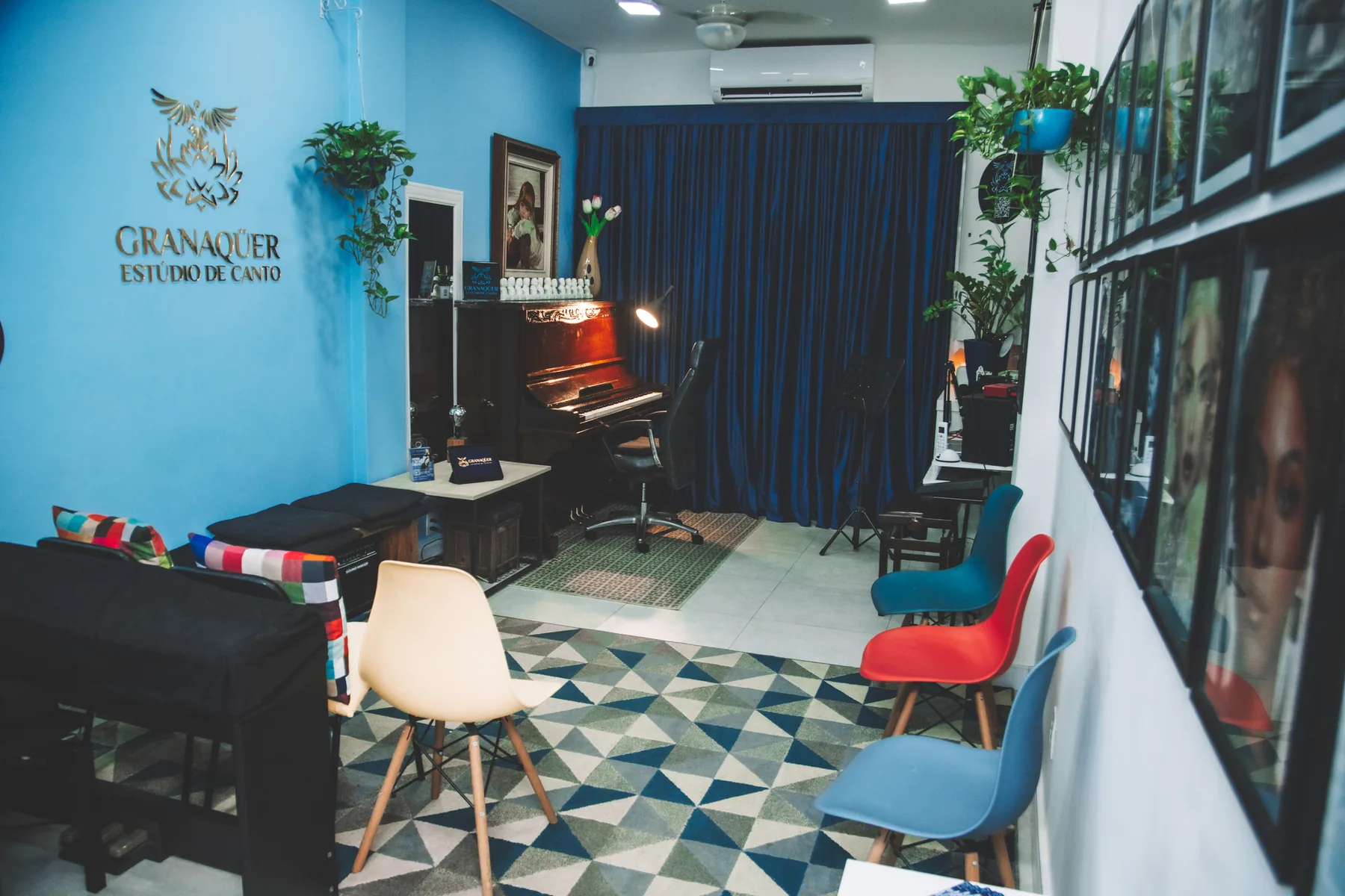 Sala de aula de canto do Granaqüer, em Botafogo: parede azul com o logotipo, piano acústico, cortina azul e cadeiras coloridas